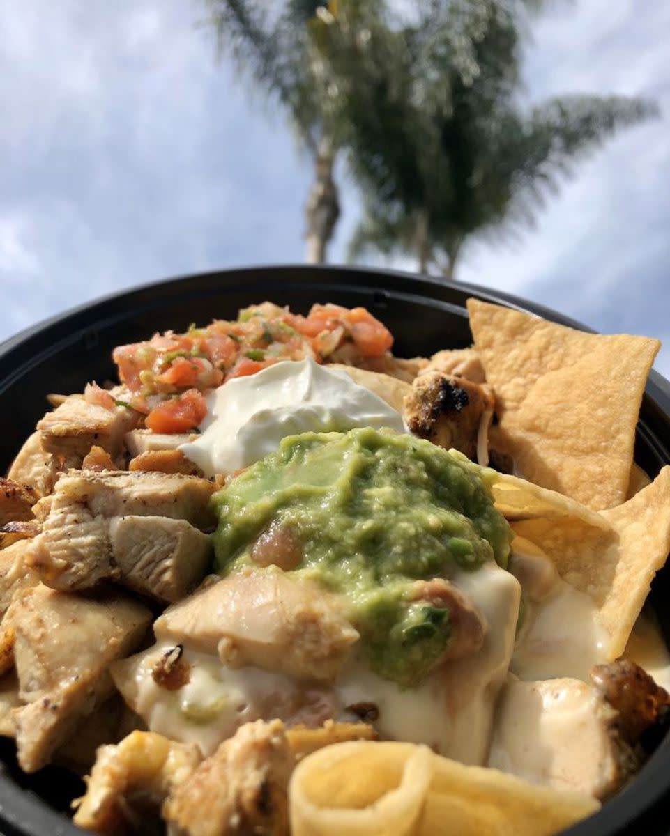 Closeup of El Pollo Loco chicken nachos with guacamole, sour cream, and salsa in a black bowl, selective focus, palm trees against a blue sky with clouds blurred in the background