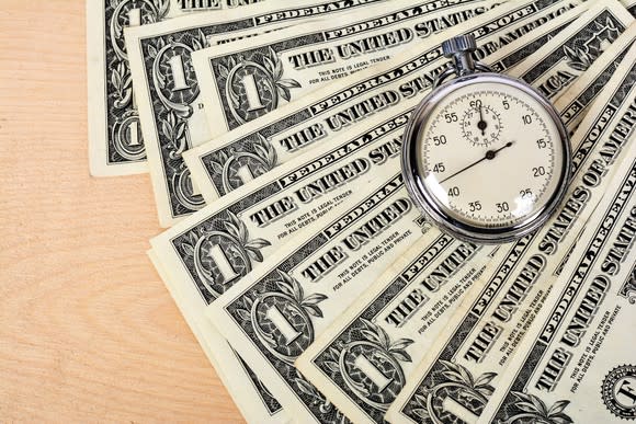 A stopwatch sits atop a pile of fanned-out dollar bills, signifying that time is money.