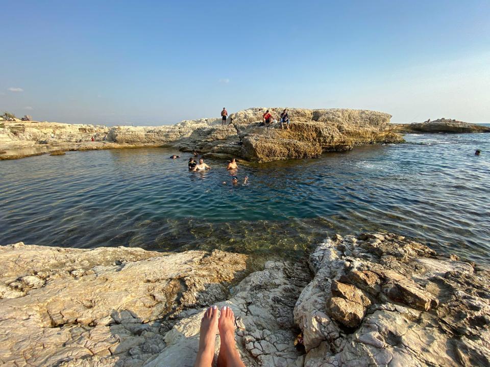 A view of a rocky beach with just someone's toes visible and people swimming in the background.