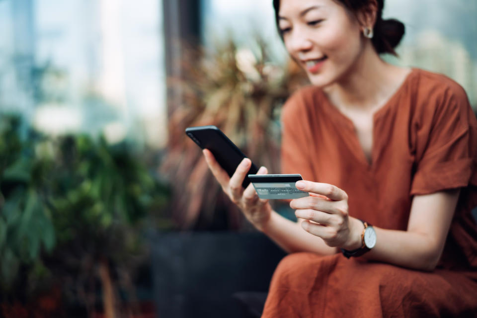 Beautiful smiling young Asian woman relaxing on deck chair in the backyard, surrounded by beautiful houseplants. Shopping online on smartphone and making mobile payment with credit card. Technology makes life so much easier. Lifestyle and technology