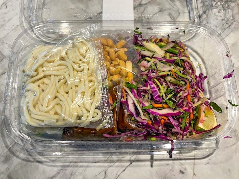 A clear plastic box containing a pack of udon noodles, peanuts, and a mixture of shredded cabbage and carrots with a lime wedge