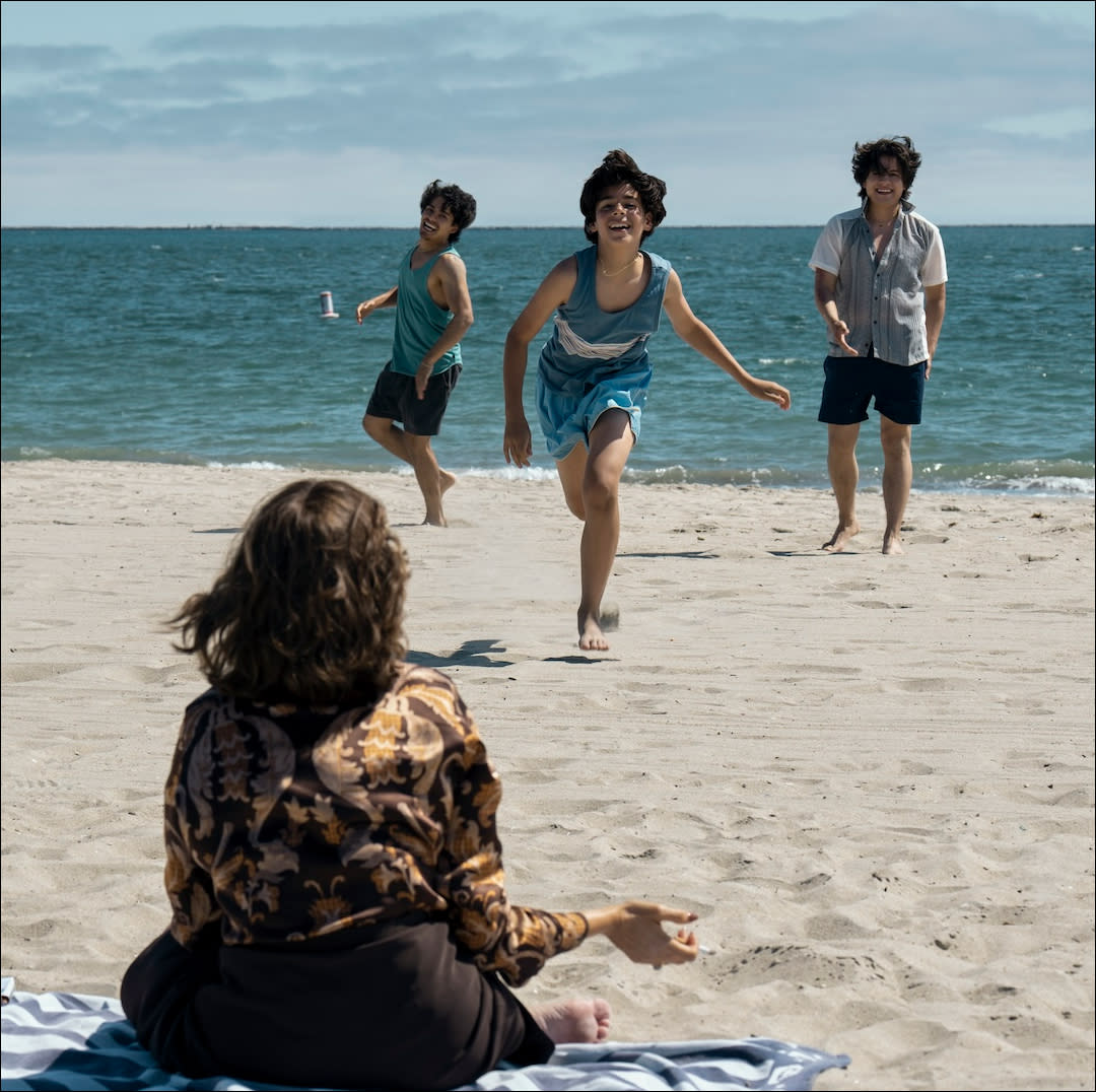  A woman (sofia vergara as griselda) sitting on a blue striped beach towel with her back to the camera faces three boys running towards her on the sand, with the sea in the background. 