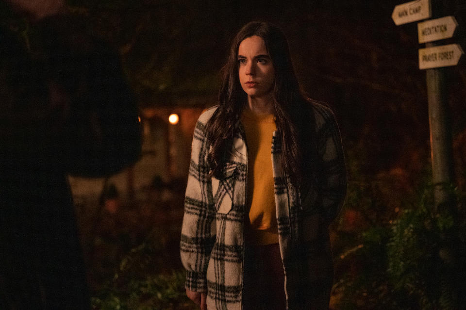 A teenage girl in a yellow shirt and plaid jacket stands alert in a compound in the woods; still from "Yellowjackets."