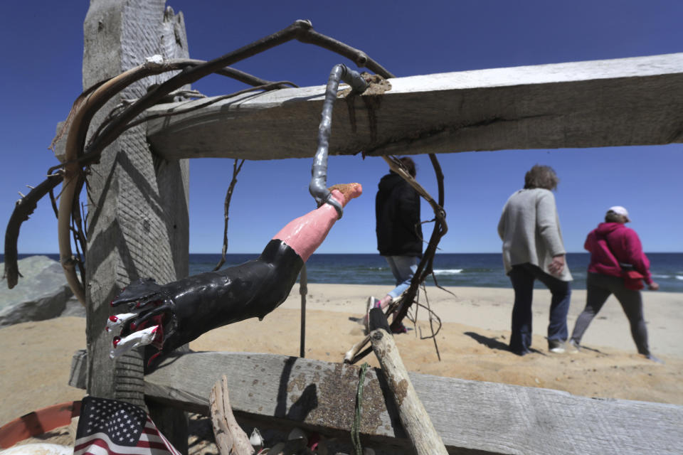 In this May 22, 2019, photo, three women pass a makeshift memorial at Newcomb Hollow Beach, where a boogie boarder was bitten by a shark and later died of his injuries the previous summer, in Wellfleet, Mass. Cape Cod beaches open this holiday weekend, just months after two shark attacks, one of which was fatal, rattled tourists, locals and officials. Some precautionary new measures, such as emergency call boxes, have yet to be installed along beaches where great whites are known to frequent. (AP Photo/Charles Krupa)