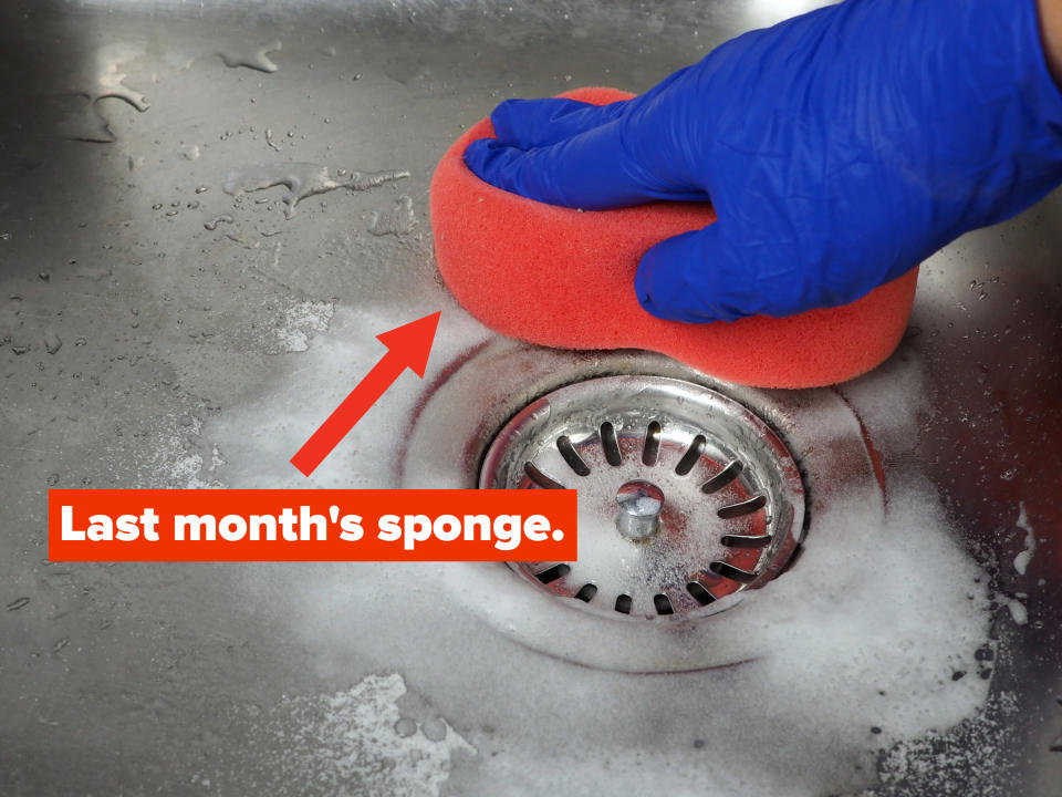 A gloved hand wipes down a soapy kitchen sink with a large kitchen sponge