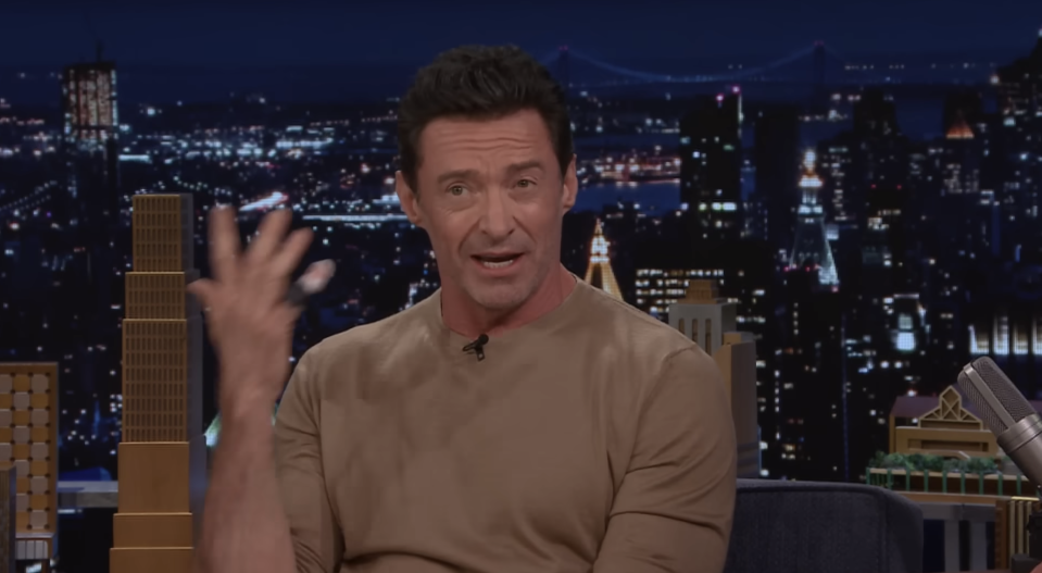 Hugh Jackman, gesturing with his hand, speaks during a night-time TV interview with a city skyline in the background