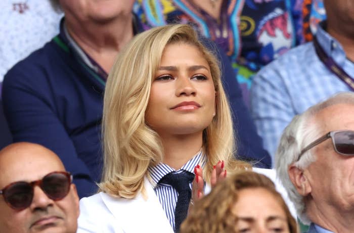 Zendaya watching an event, wearing a striped shirt, tie, and white jacket, with blonde hair styled in soft waves