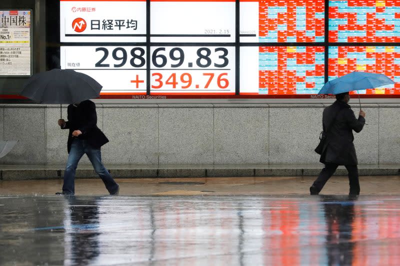 Men holding umbrellas walk in front of an electric board showing Nikkei index at a brokerage in Tokyo