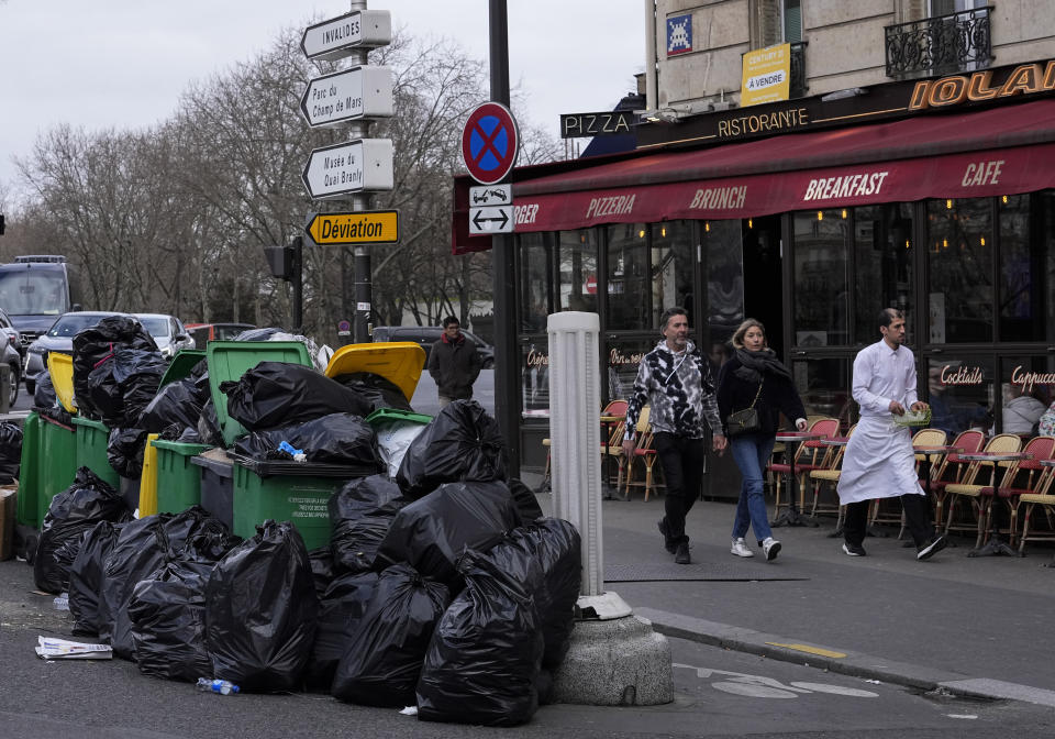 People walk past not collected garbage cans next to the Senate in Paris, Sunday, March 12, 2023. A contentious bill that would raise the retirement age in France from 62 to 64 got a push forward with the Senate's adoption of the measure amid strikes, protests and uncollected garbage piling higher by the day. (AP Photo/Michel Euler)