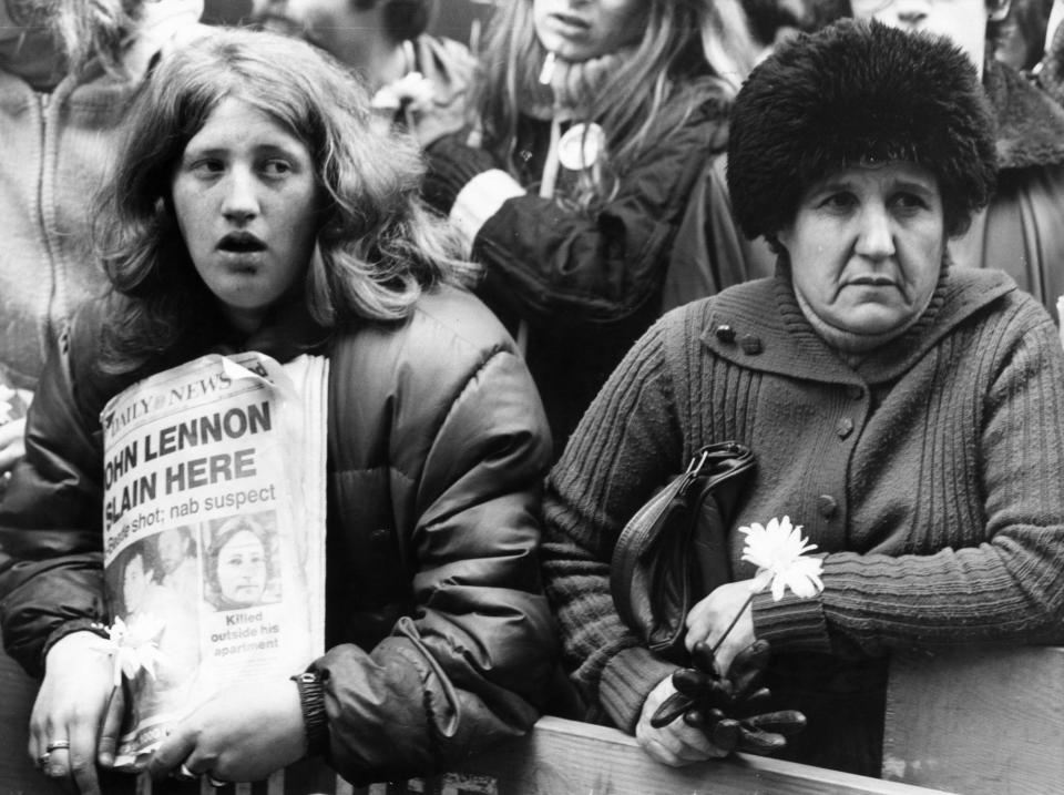 Two women are in a crowded place. One holds a newspaper with the headline 