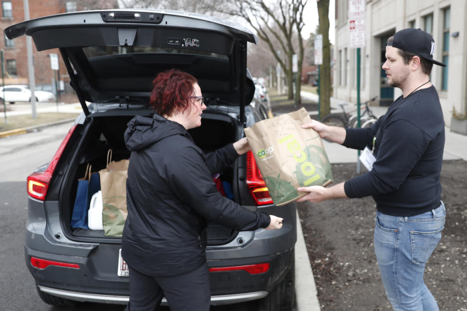 Sugar Beets Food Co-Op employee Jamie Melendy, right, gives curb service to Lola Wright in the Village of Oak Park, Ill., Friday, March 20, 2020. There are at least three confirmed cases of COVID-19 in Oak Park, just nine miles from downtown Chicago, where the mayor has ordered residents to shelter in place. With so few tests available, surely there are others, says Tom Powers, spokesman for the village of about 52,000 in a metropolitan area with millions. (AP Photo/Charles Rex Arbogast)