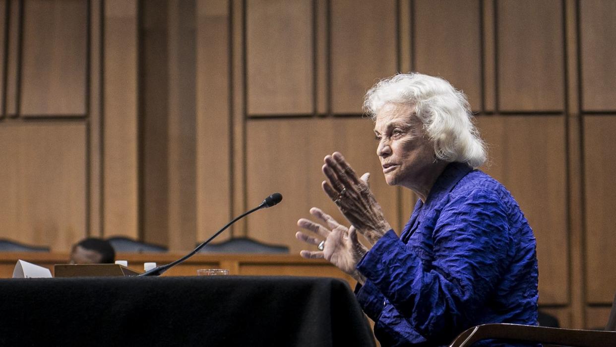 sandra day o'connor sits in a chair at a table and speaks into a microphone, she wears a blue jacket and has her hands raised in front of her