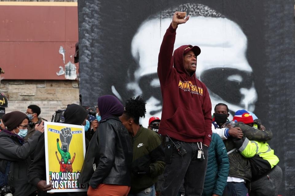 A black man in a hooded sweatshirt holds his right fist in the air. Other people are hugging behind him.