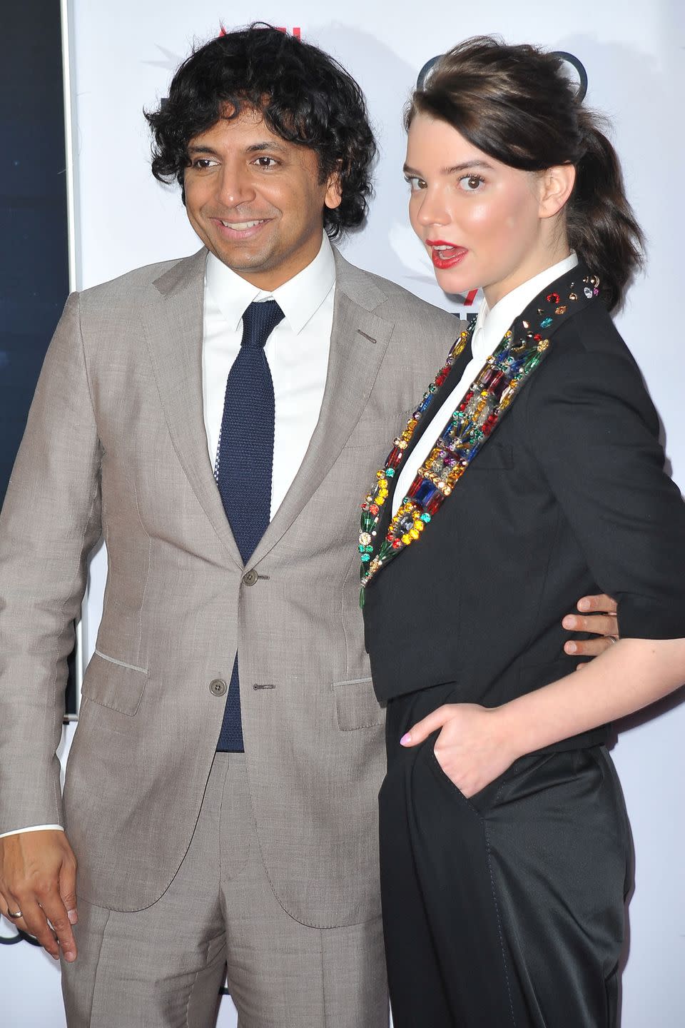 m night shyamalan and anya taylor joy pose for photos while standing together, he wears a gray suit with a white collared shirt and navy tie and smiles as he looks to the left, she wears a black suit with a large beaded multicolor necklace and looks over her shoulder at the camera