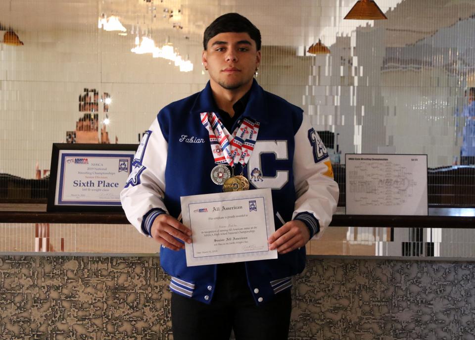 Fabian Padilla showcases his senior wrestling awards in 2019. On the left is his sixth-place finish at Virginia Beach. On his right is his State Championship bracket. He holds his All-American wrestler certificate and wears his three state wrestling medals around his neck.