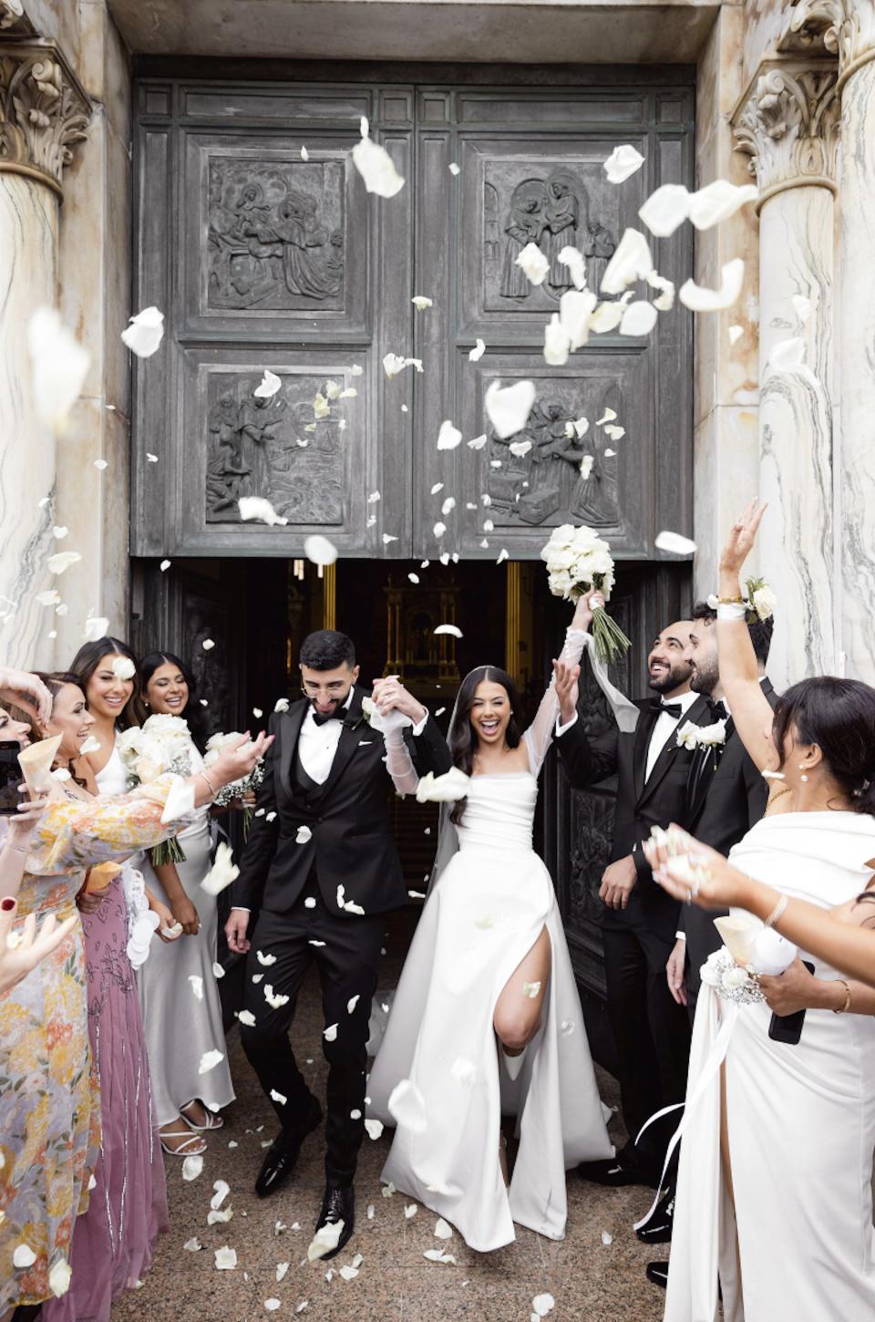 A bride and groom raise their hands in celebration as their wedding guests throw flower petals on them.
