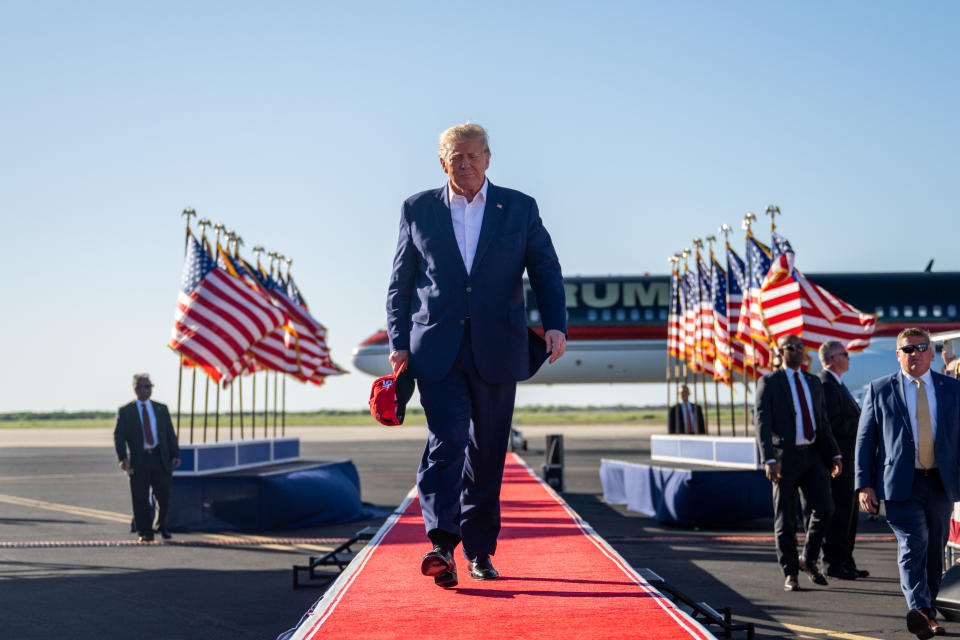 Former President Donald Trump walks down a red carpet outside an airport, with his private plane and rows of American flags in the background.