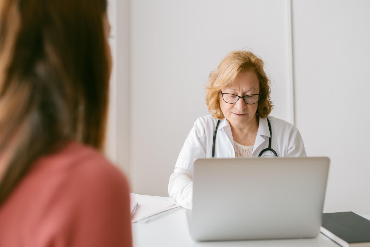 A girl sits across from a gynecologist, who is looking down at a laptop computer.