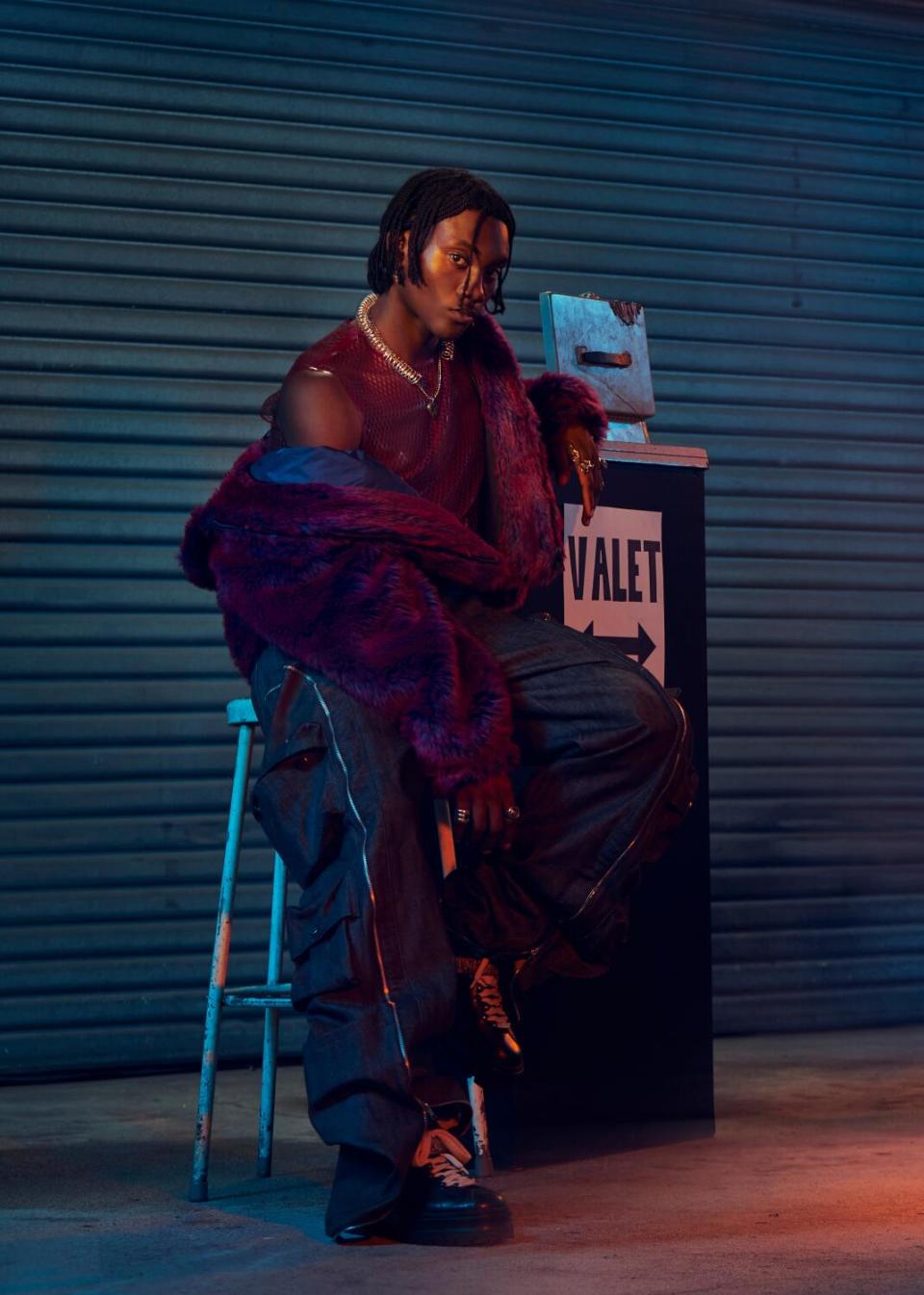 A model sits beside a "Valet" sign, his Givenchy jacket falling off his shoulders.