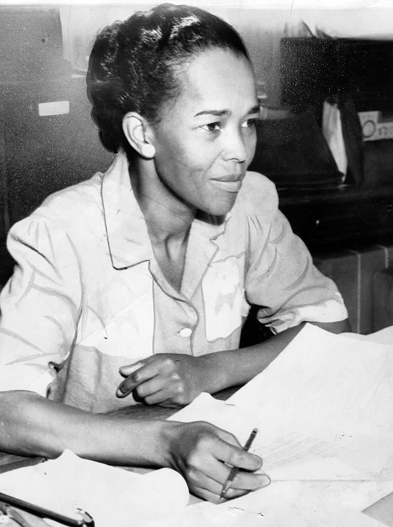 ella baker sits a table with several papers in front of her and holds a pen in one hand, she wears a collared shirt and looks right