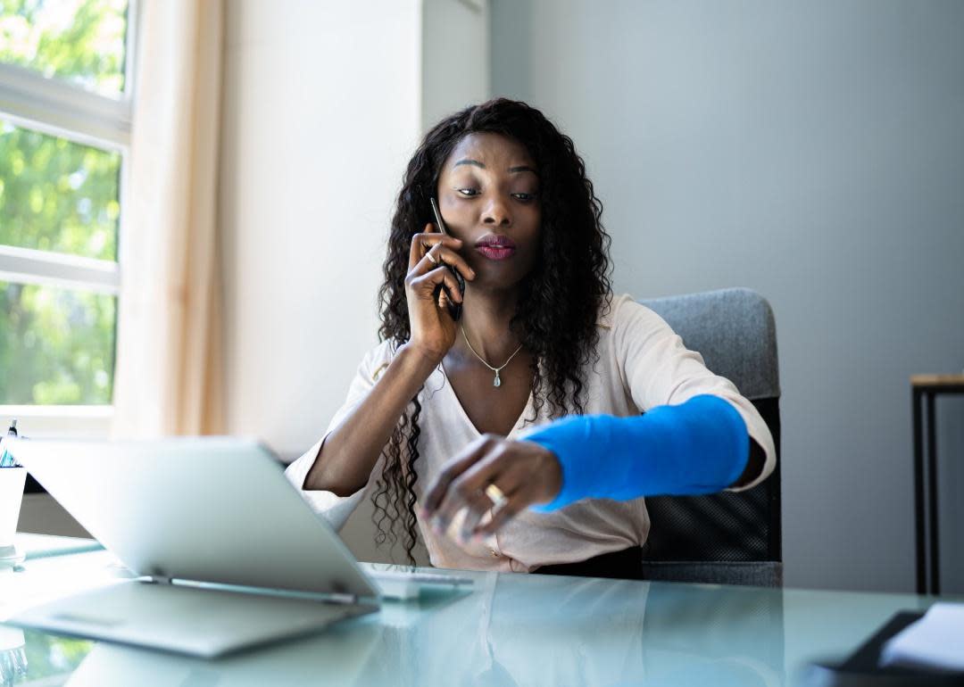 A woman at a desk on the phone with a blue cast on her arm.