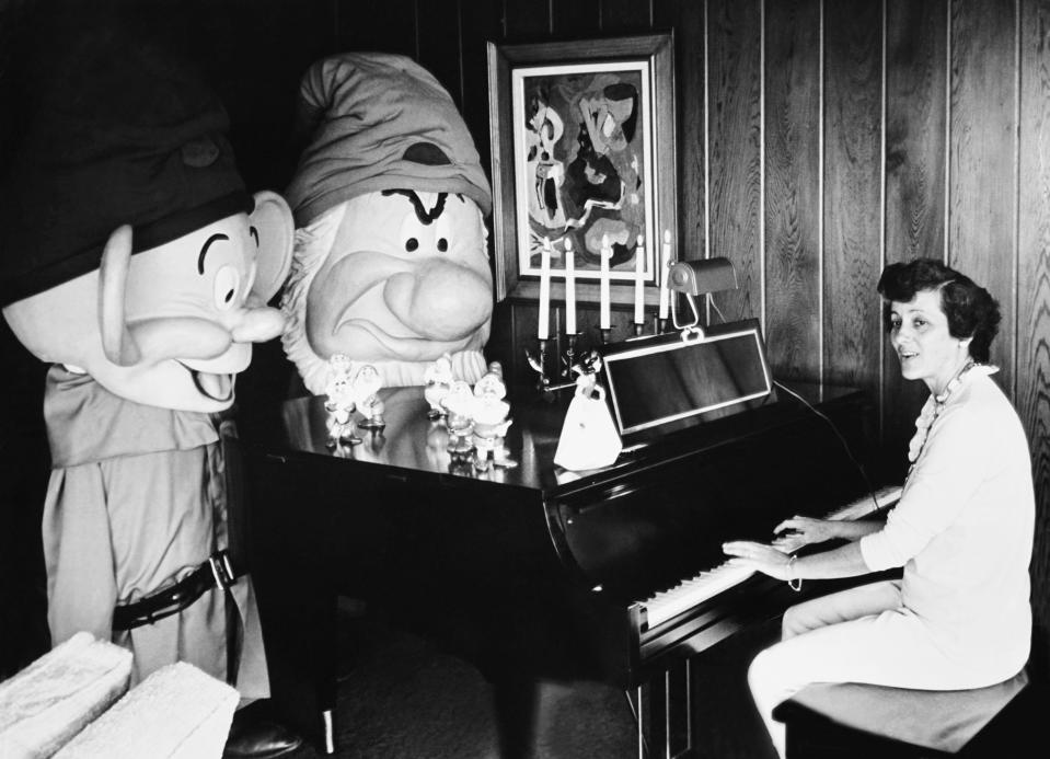 A woman playing piano with two people dressed as Snow White's dwarfs standing next to the piano