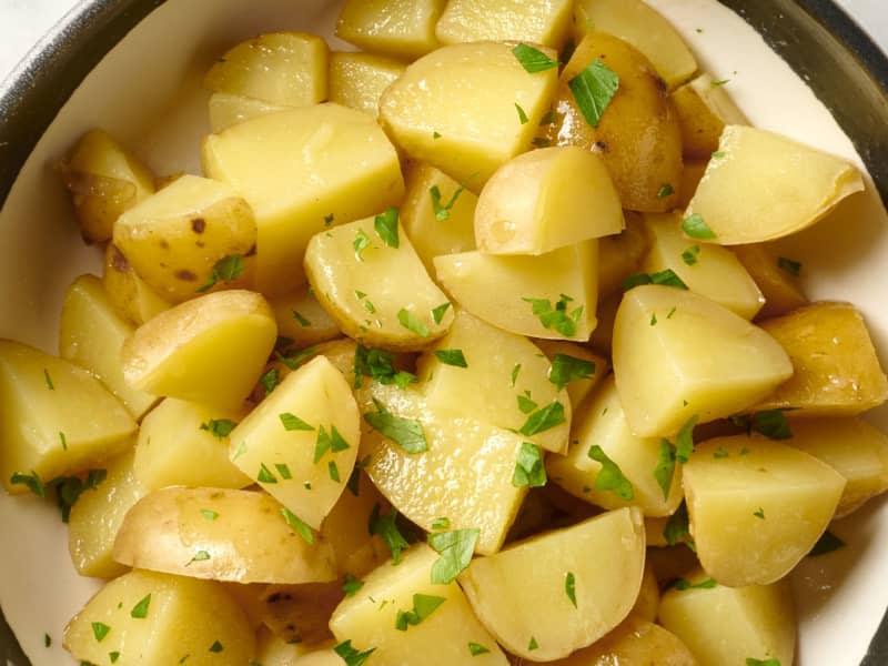 Overhead view of boiled potatoes in a white bowl with a brown rim, topped with herbs.