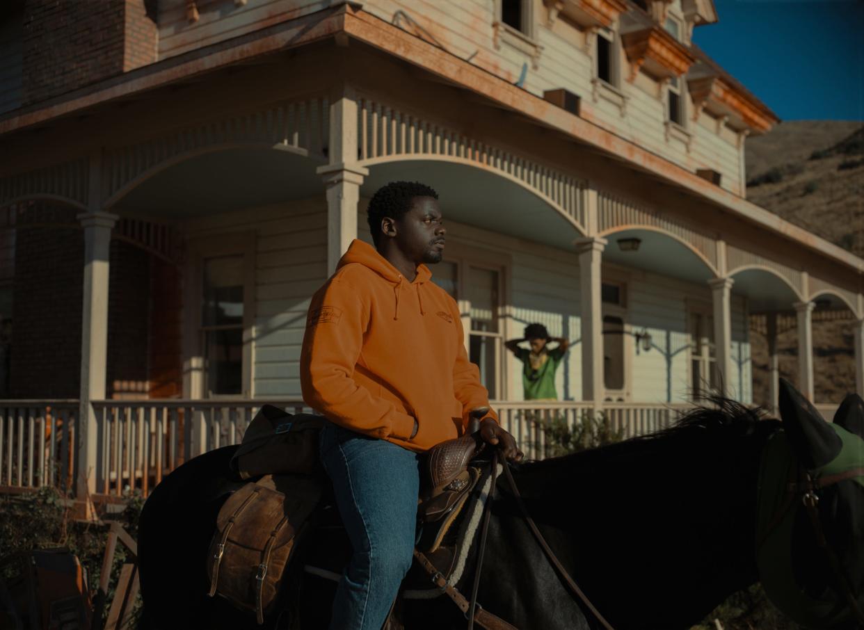OJ sits astride a horse in an orange hoodie, while Emerald stands on the porch of their home behind him in a green jersey.