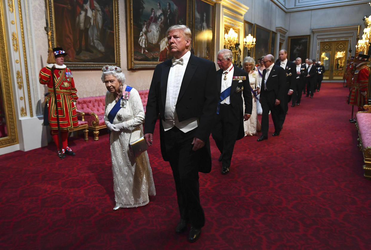 Queen Elizabeth II, looking bowed with age, and President Donald Trump, standing tall, lead a long file of dignitaries, with a Beefeater standing to attention in the background.