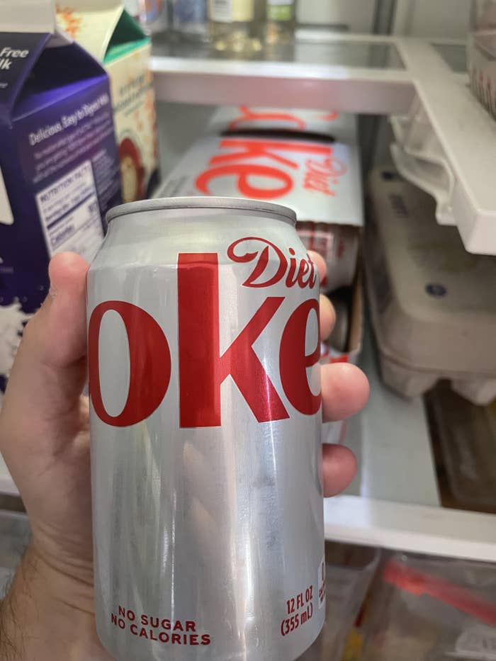 A hand holding a can of Diet Coke in front of an open fridge with a 12-pack of Diet Coke in it