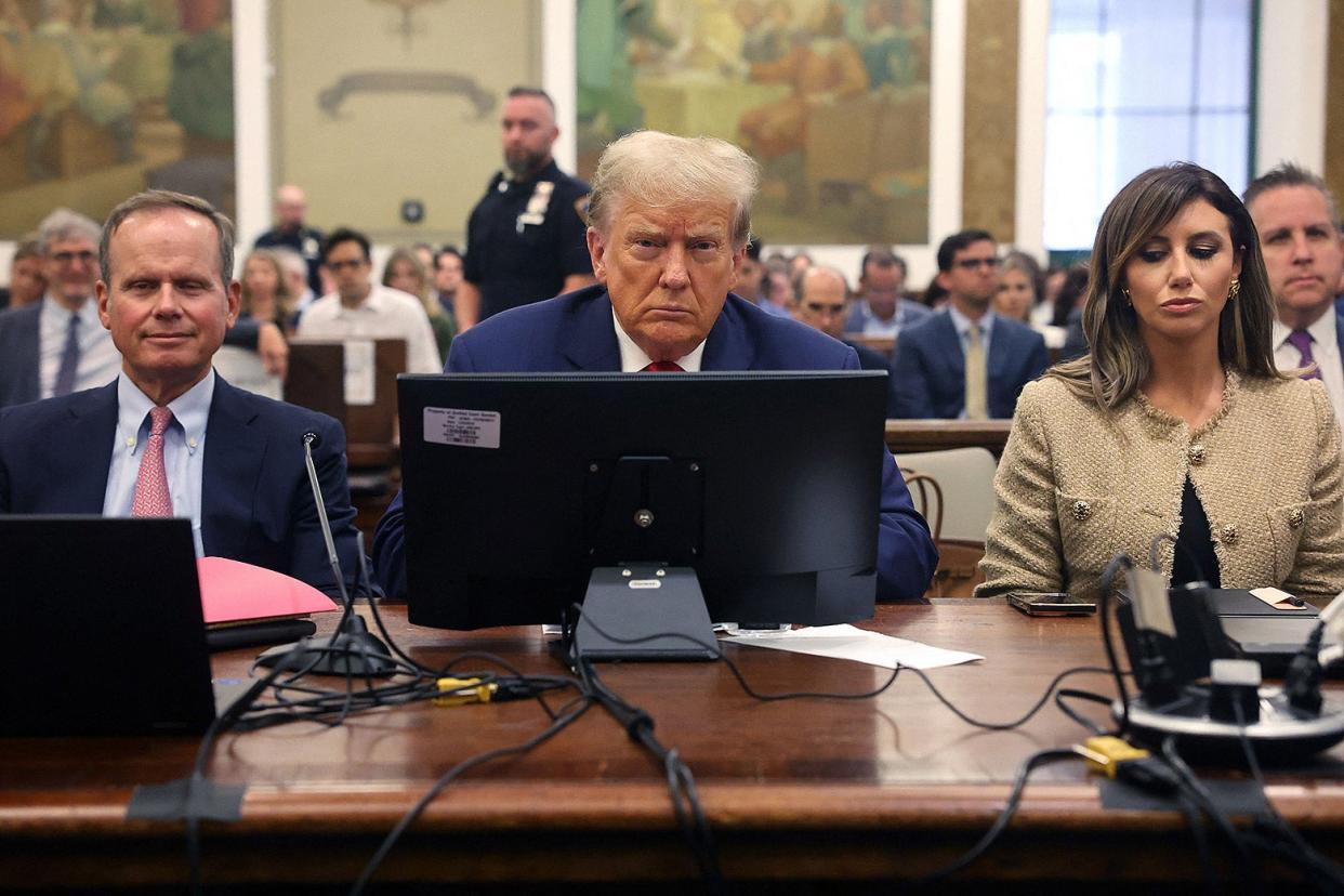 Trump frowning at the defendant's table with a computer in front of him and his attorneys by his side.
