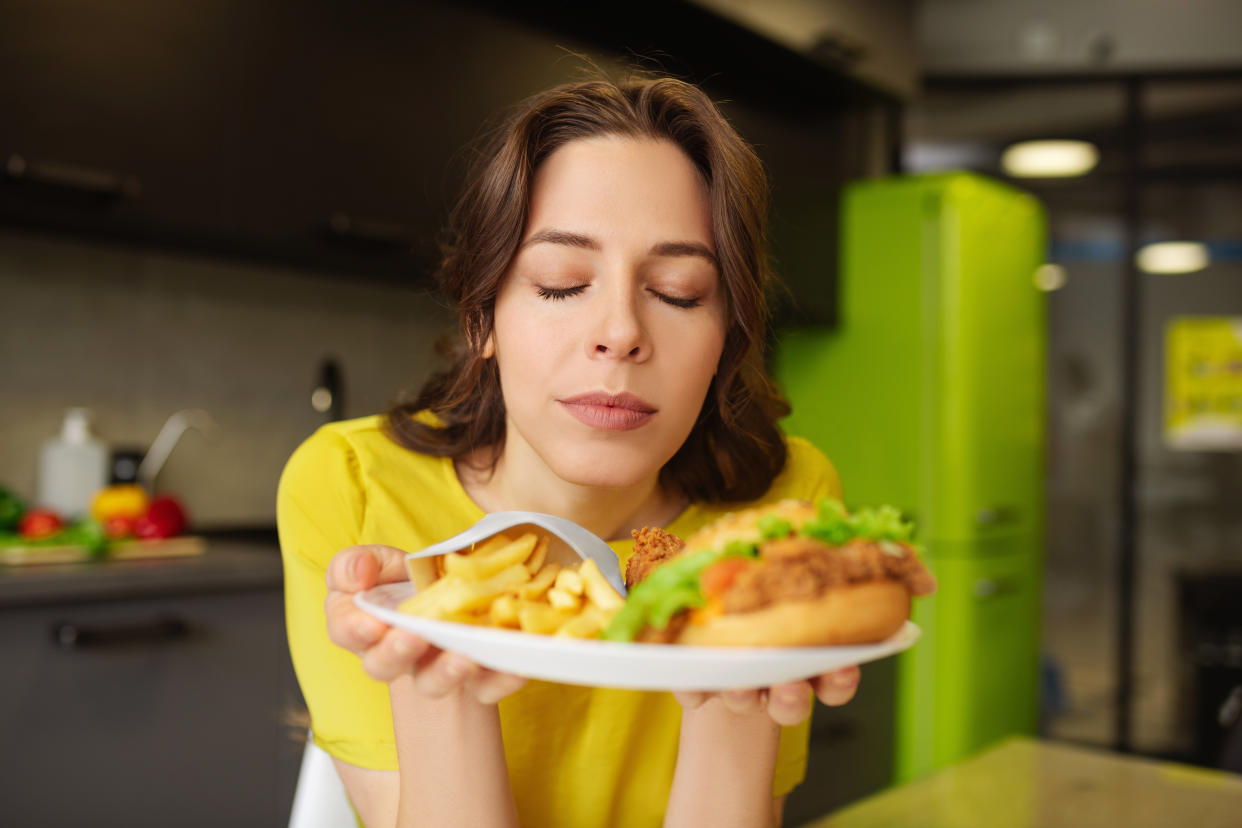 Temptation. Happy long-haired woman with closed eyes sitting in the kitchen at the table with a plate with a burger french fries