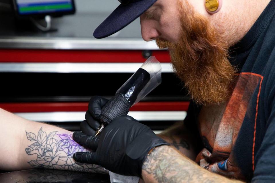 Using a tattoo gun, Jonathan Pope of Lowell and owner of From The Ashes Tattoo Studio, tattoos Sarah Mente’s arm, adding to another tattoo he inked.