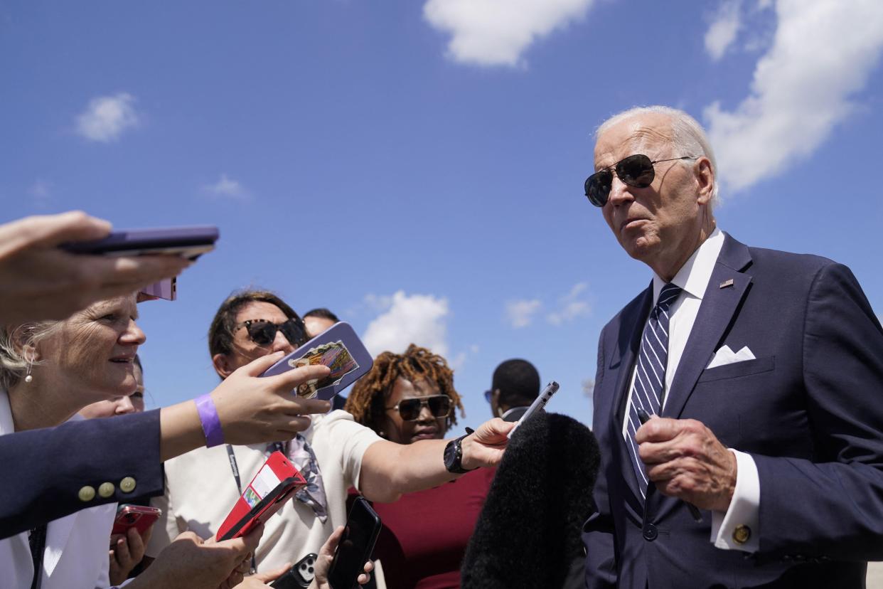 President Biden, wearing a navy suit, white shirt, and aviator sunglasses, speaks to reporters holding out phones and voice recorders.