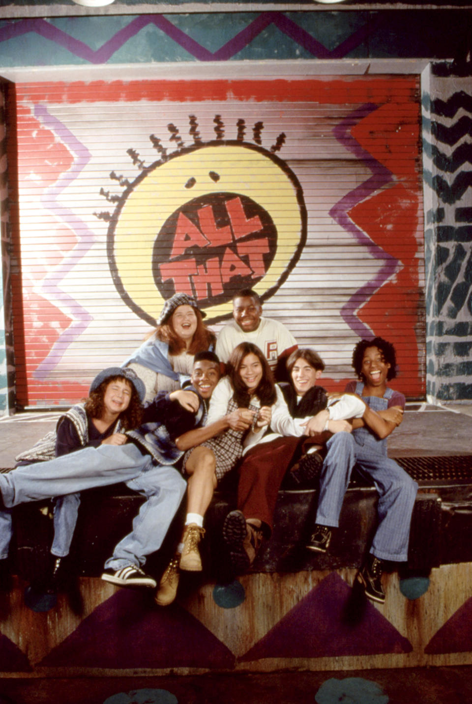 Cast of "All That" posing in front of show's logo, in casual 90s attire, smiling