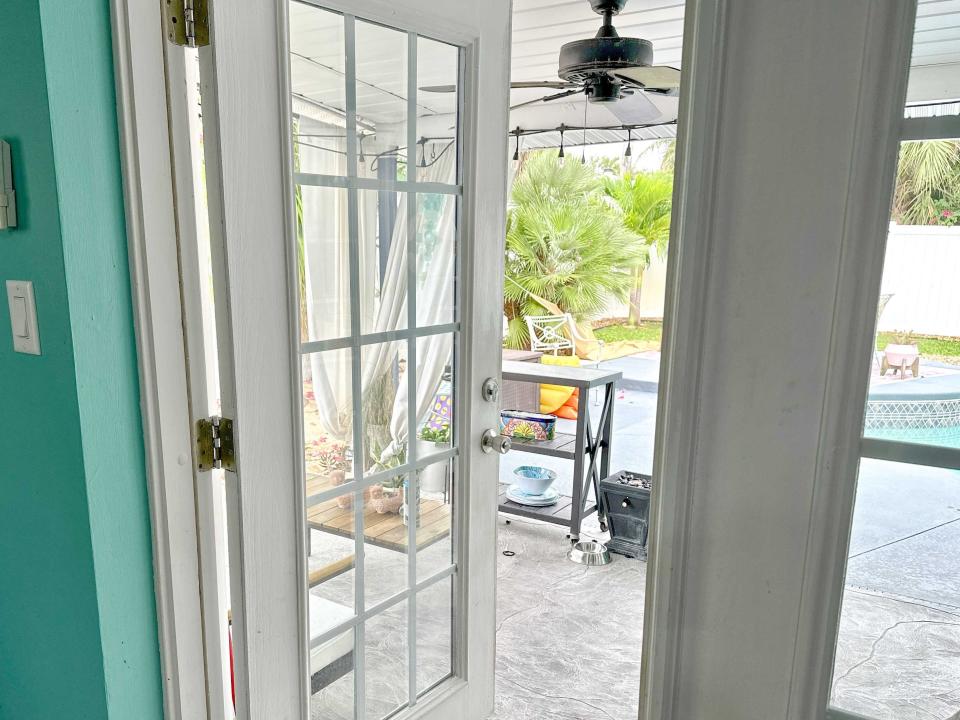 An open white door leading to a patio with a stone ground, a table, and a backyard with a pool, palm trees, and white chairs