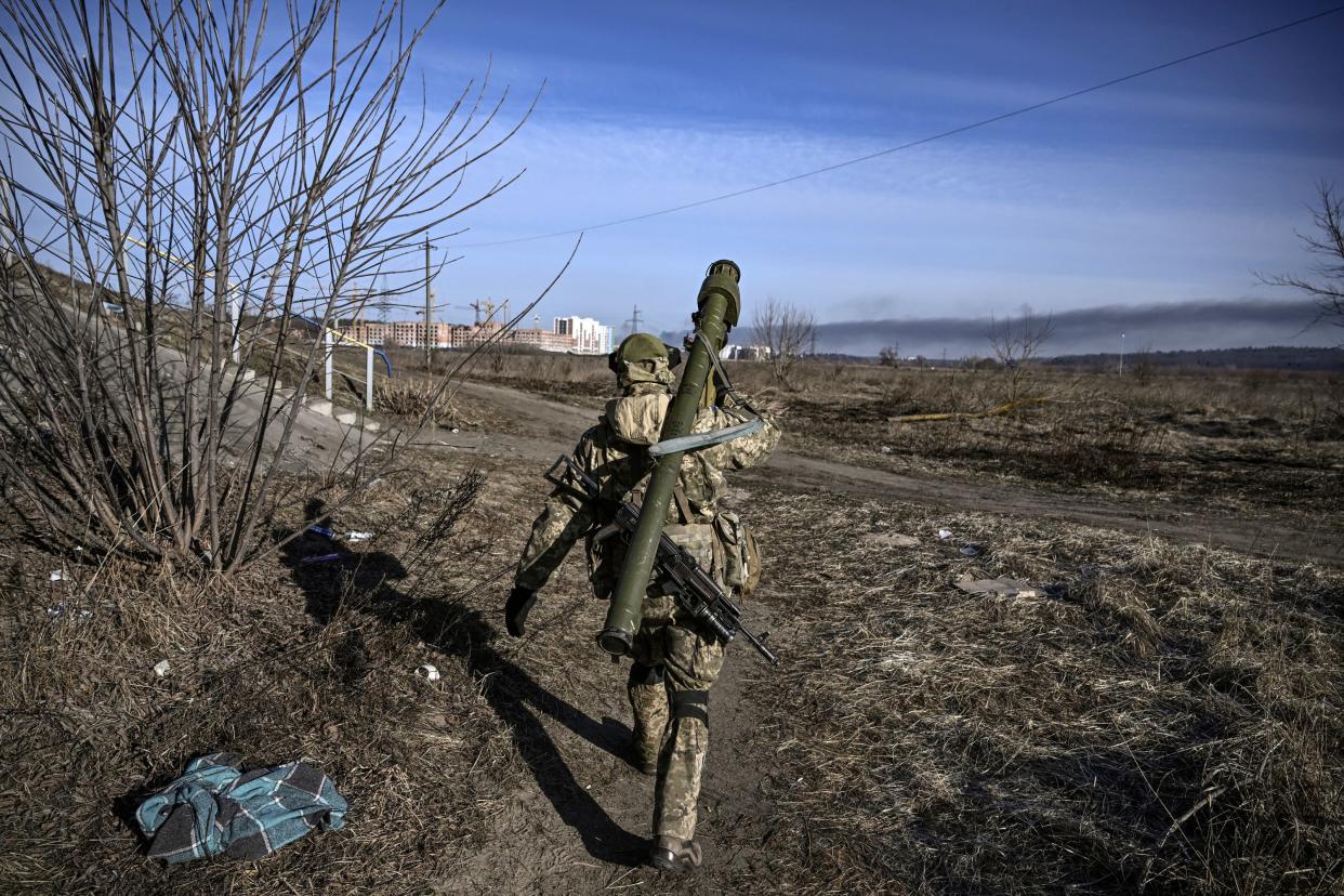 A serviceman in fatigues and carrying weaponry on their back walks along a path in a barren field toward buildings in the far distance.  