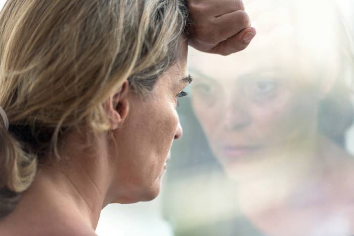 A woman with light hair leans her forehead against a glass window, with her reflection visible. Her expression appears contemplative or sad