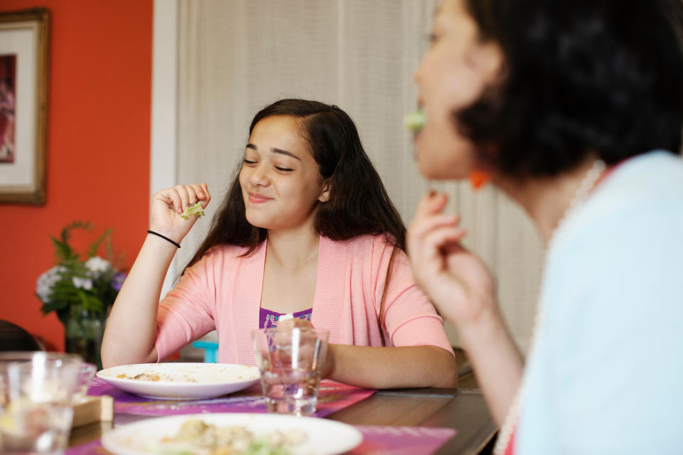 Two people are seated at a dining table, enjoying their meal. The person in focus has long hair and is smiling as they eat. The other is slightly blurred