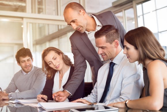 A man stands over a team of professionals, reviewing documents.