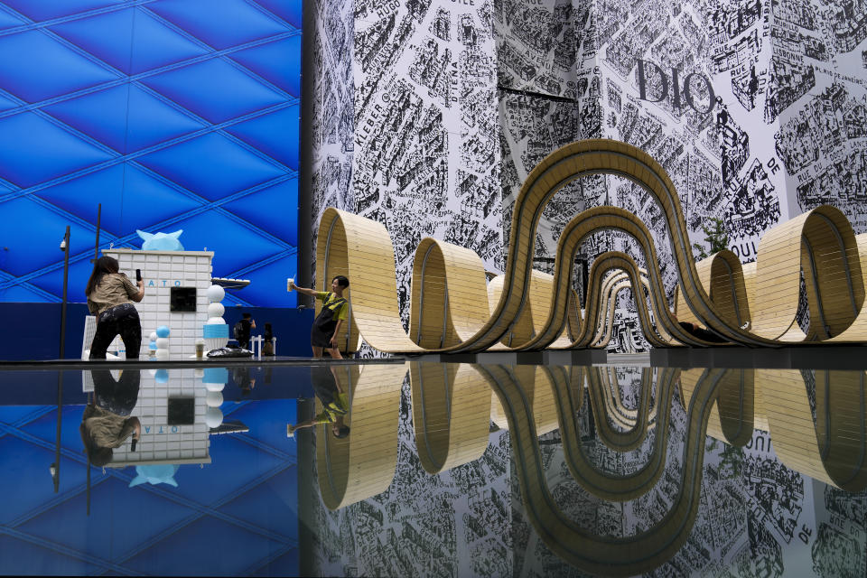 A woman holding a cup of coffee poses for a souvenir photo with an art installation reflected at an outdoor luxury shopping mall in Beijing, Wednesday, June 5, 2024. (AP Photo/Andy Wong)