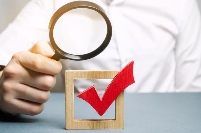 A person looking at a wooden sculpture of a checkmark in a box through a magnifying glass.