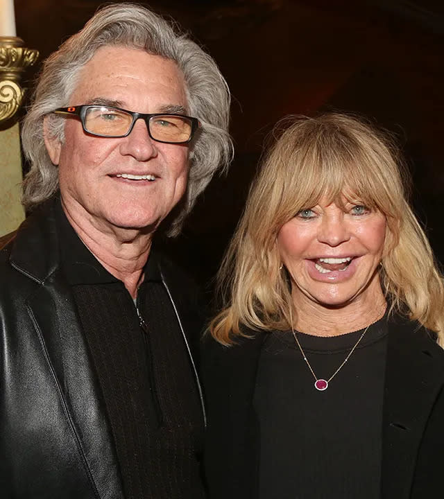 Kurt Russell in a sweater and black leather jackets smiles next to a happy Goldie Hawn in a black sweater and jacket and pink-stoned necklace