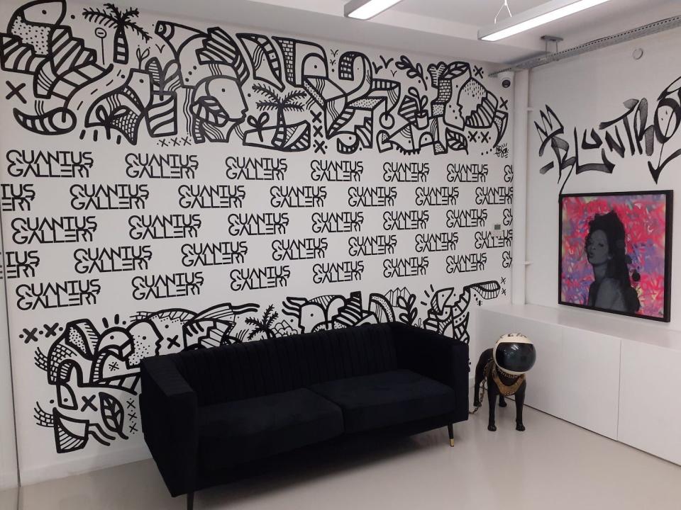 A black sofa sits against a white wall., featuring the words Quantus Gallery printed multiple times in a repeated pattern. Next to the sofa is a sculpture featuring a dog wearing a space helmet, and a bright pink picture featuring a woman.