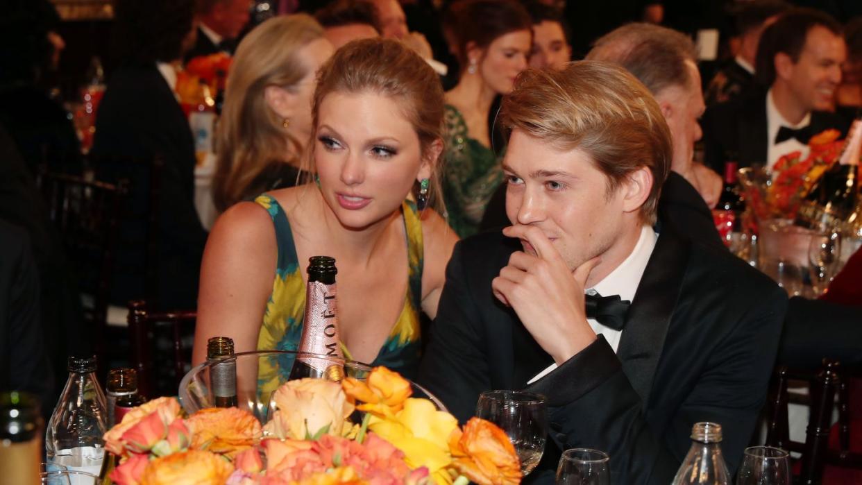 taylor swift and joe alwyn sit at a table with yellow and orange flowers and several drinks on it, she wears a sleeveless blue and orange dress, he wears a black tuxedo and bowtie, they look to the left