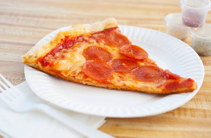 A slice of pepperoni pizza on a white paper plate sits on a wooden table, accompanied by plastic utensils and condiment cups