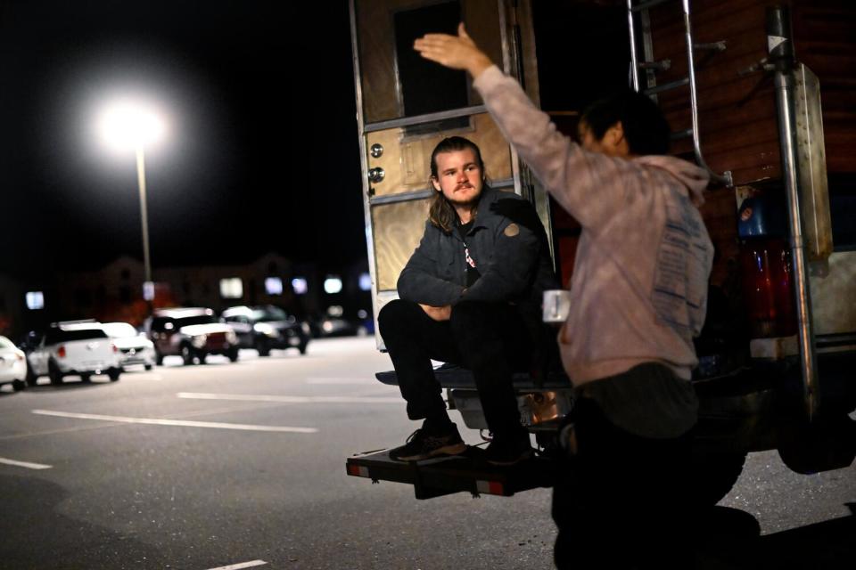 Two people, one seated on a vehicle's hitch rack talk in a parking lot at night.