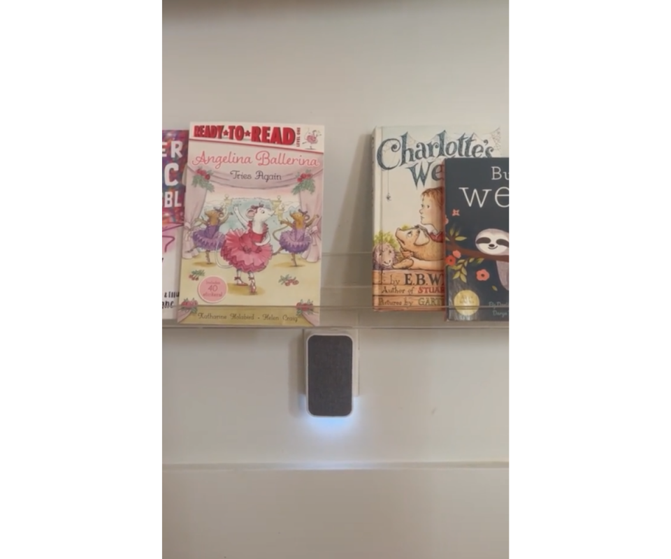 A grey rectangular doorbell receiver sits underneath a children's bookshelf on a white wall.