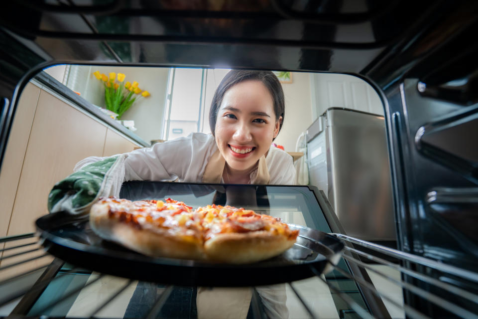 Happy Young Asian Woman looking at camera while cooking and put Pizza in Oven at kitchen at Home . Italian food, cooking, and people lifestyle concept.