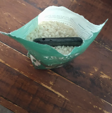 A packet of rice, opened and placed upright on a wooden surface, with a black electronic device submerged in the rice grains